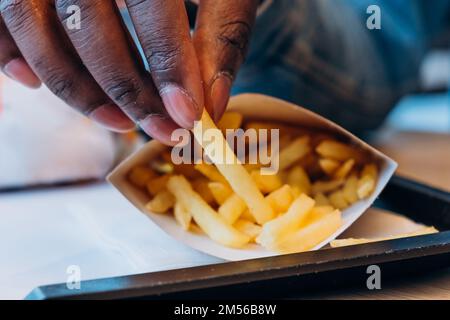 Ein junger afroamerikanischer Mann nimmt leckere frische pommes aus dem Papierbehälter an den Tisch im modernen Café, ganz aus der Nähe Stockfoto