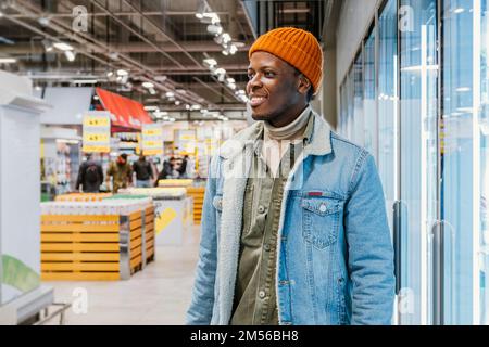 Fröhlicher junger afroamerikanischer Kerl in warmer Denim-Jacke steht in der Nähe von Kühlschränken in der modernen Supermarktabteilung Stockfoto