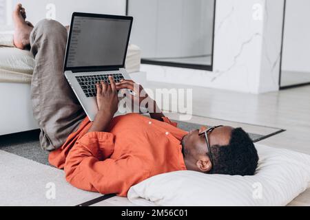 Barfuß afroamerikanischer Freiberufler in orangefarbenem Hemd arbeitet auf einem modernen Laptop, der auf dem Boden in der Nähe einer bequemen Bettseitenansicht liegt Stockfoto