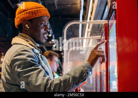 Ein positiver afroamerikanischer Mann mit orangefarbenem Strickhut und kabellosen Kopfhörern bestellt Essen über einen Selbstbedienungskioské im Café Stockfoto