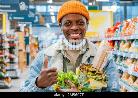 Lächelnder afroamerikanischer Mann in warmem orangefarbenen Hut mit Einwegmaske zeigt Daumen nach oben mit Salaten in der Supermarkt-Abteilung Stockfoto