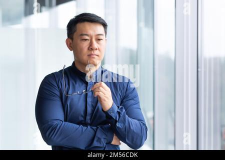 Porträt eines seriösen jungen asiatischen Geschäftsmanns, Anwalt. Er steht durchdacht im Büro am Fenster, hält die Brille in den Händen und schaut zur Seite. Stockfoto