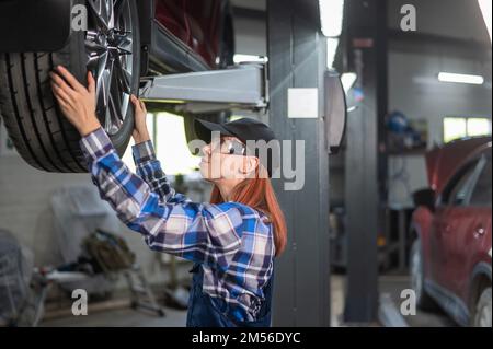 Weibliche Mechanikerin, die den Reifen des Fahrzeugs auf der Hebebühne justiert. Ein Mädchen bei der Arbeit eines Mannes. Stockfoto