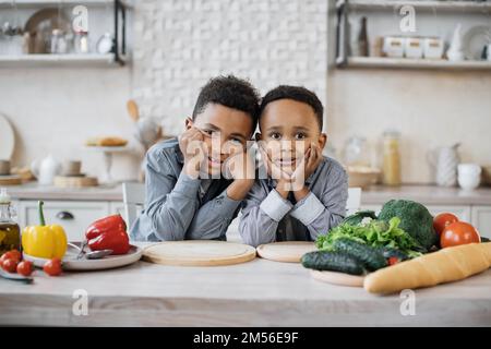 Lächelnde kleine süße afrikanische Brüder in legerer Kleidung, die am Tisch sitzen und die Wangen berühren, die mit Salat gefüllt sind. Wunderschöne afroamerikanische Kinder bereiten gesundes Essen in der Küche zu. Stockfoto