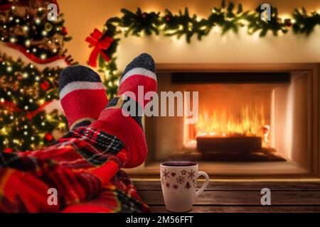 Entspannen Sie sich vor dem gemütlichen Kamin mit erhobenen Füßen und tragen Sie Weihnachtssocken auf dem Couchtisch. Weihnachtsdekoration, warmes orangefarbenes Feuer und dunkle Stimmung im Innenraum Stockfoto