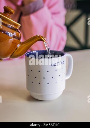 Heißer Tee in Teekanne und Tasse mit Dampf auf einem Tisch. Frau gießt Tee aus brauner Teekanne in eine Tasse. Der Prozess der Teezubereitung, bei dem heißes Wasser aus dem K ausgeschüttet wird Stockfoto