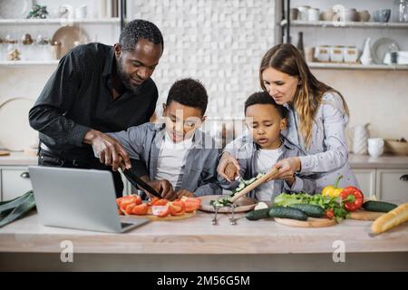 Fröhliche afrikanische süße kleine Söhne und ihre jungen Eltern, die zusammen einen Online-Kochkurs auf einem Laptop haben, mit frischem Gemüse und Salat, Essen in der Küche zu Hause. Stockfoto