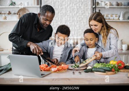 Fröhliche afrikanische süße kleine Söhne und ihre jungen Eltern, die zusammen einen Online-Kochkurs auf einem Laptop haben, mit frischem Gemüse und Salat, Essen in der Küche zu Hause. Stockfoto