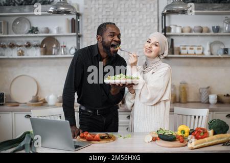 Fröhliches, multinationales Ehepaar, das ein Notebook benutzte, während es gesundes Essen in der Küche kochte, junge muslimische Frau im Hijab fütterte ihren Mann mit einer Gabel mit Salat aus frischem Gemüse. Stockfoto