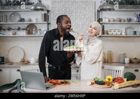 Fröhliches, multinationales Ehepaar, das ein Notebook benutzte, während es gesundes Essen in der Küche kochte, junge muslimische Frau im Hijab fütterte ihren Mann mit einer Gabel mit Salat aus frischem Gemüse. Stockfoto