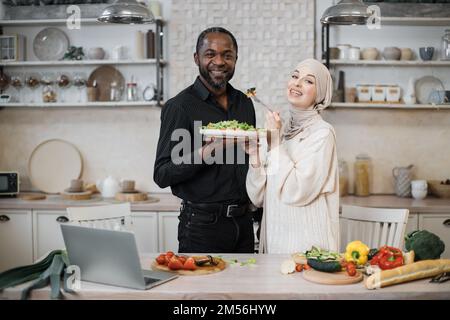 Fröhliches, multinationales Ehepaar, das ein Notebook benutzte, während es gesundes Essen in der Küche kochte, junge muslimische Frau im Hijab fütterte ihren Mann mit einer Gabel mit Salat aus frischem Gemüse. Stockfoto