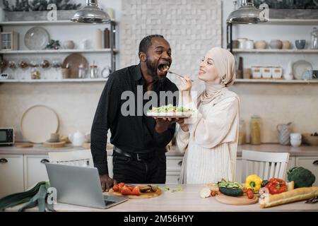 Fröhliches, multinationales Ehepaar, das ein Notebook benutzte, während es gesundes Essen in der Küche kochte, junge muslimische Frau im Hijab fütterte ihren Mann mit einer Gabel mit Salat aus frischem Gemüse. Stockfoto