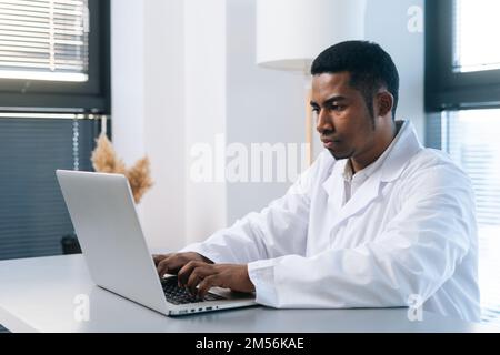 Porträt eines schweren schwarzen männlichen Arztes, der weiße Kitteluniform trägt und an einem Laptop-Computer arbeitet, der am Schreibtisch in einer leichten medizinischen Klinik sitzt. Stockfoto