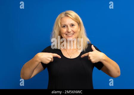 Porträt einer charmanten, glücklichen Frau in einem schwarzen T-Shirt, die lächelt und auf sich selbst zeigt, stolz auf ihren eigenen Erfolg ist und mit ihren Leistungen prahlt Stockfoto