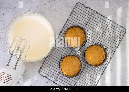 Käsekuchen-Rezept. Drei kleine runde Kuchenpfannen (Dose, Schimmel) mit zerdrückten Crackern und Käsekuchen-Teig in einer Schüssel, flach liegend Stockfoto