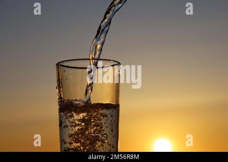 Sauberes Wasser, das auf dem Hintergrund des Sonnenuntergangs in das Trinkglas fließt. Konzept von Gesundheit und Frische, Durst, Wasserreinigung Stockfoto