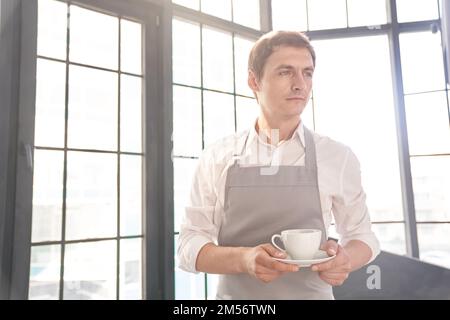 Ein männlicher Kellner in einer grauen Schürze hält eine Tasse Kaffee. Der Barista serviert eine Tasse heißen Kaffee vor dem Hintergrund eines großen Fensters mit Platz zum Kopieren. Hochwertiges Foto Stockfoto