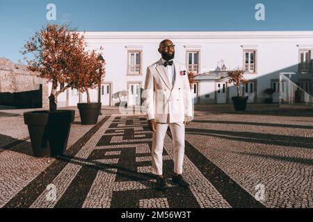 Ein eleganter schwarzer Mann mit Glatze, elegant gekleidet in einem weißen Anzug, einer Fliege und einer Brille steht auf der portugiesischen Pflastersteinpflasterei neben großen Pflanzen im Freien Stockfoto