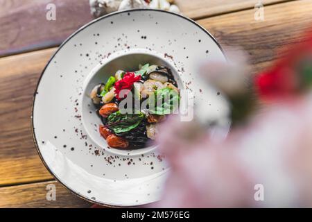 Meeresfrüchte- und Nudelkonzept. Garnelen serviert mit schwarzer Tagliolini-Pasta auf einem weißen Tisch. Unscharfe Blumen im Vordergrund. Hochwertiges Foto Stockfoto