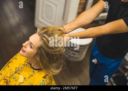 Friseurin berührt langes blondes Haar seiner jungen, erwachsenen Klientin. Draufsicht. Speicherplatz kopieren. Unscharfer Hintergrund. Hochwertiges Foto Stockfoto