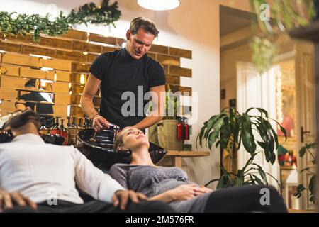 Zwei Personen sitzen auf Shampoo-Stühlen im Friseursalon. Schwangere Frau, deren blondes Haar von einem professionellen Friseur gewaschen wird. Hochwertiges Foto Stockfoto