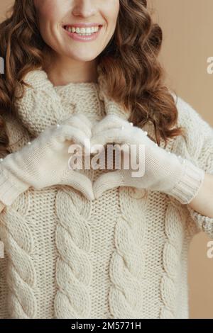 Hallo Winter. Nahaufnahme einer glücklichen Frau mittleren Alters in beigefarbenem Pullover, Fäustlingen und Hut mit herzförmigen Händen vor beigefarbenem Hintergrund. Stockfoto