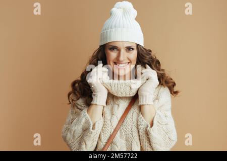 Hallo Winter. Porträt einer glücklichen, eleganten Frau in beigefarbenem Pullover, Fäustlingen und Hut vor beigefarbenem Hintergrund. Stockfoto