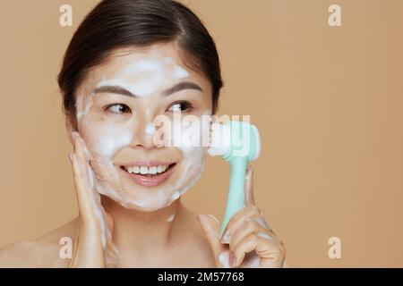 Junge Frau mit Massagegerät, Waschgesicht auf beigefarbenem Hintergrund. Stockfoto