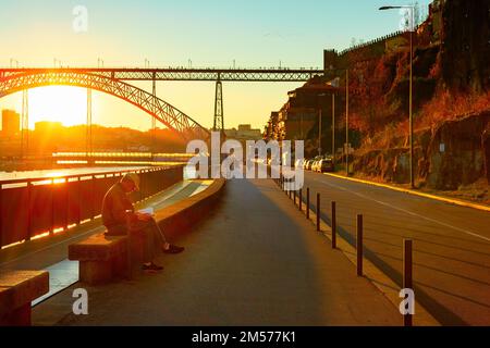 PORTO, PORTUGAL - 21. NOVEMBER 2021: Mann liest Buch über den Ufer bei Sonnenuntergang, Blick auf die Dom Luis Brücke, Porto, Portugal Stockfoto