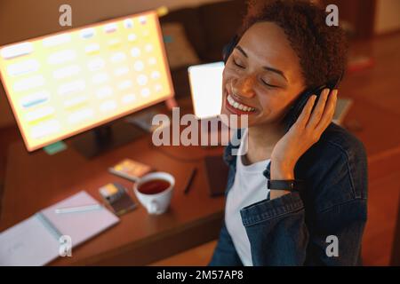 Eine lächelnde Grafikdesignerin hört in der Pause im Heimbüro gern Musik über Kopfhörer Stockfoto
