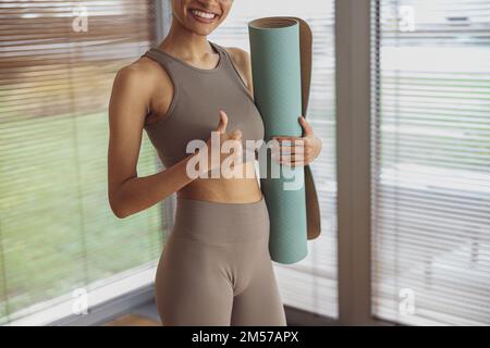 Frau hält gerolltes Yoga oder Fitnessmatte und zeigt nach dem Training zu Hause den Daumen nach oben Stockfoto