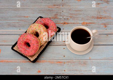 Drei Donuts in pinkfarbener und weißer Glasur mit Kokosnussflocken und eine Tasse heißem schwarzen Kaffee auf einem Holztisch. Draufsicht, flach liegend. Stockfoto