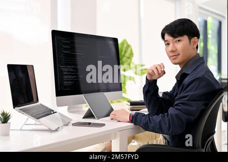 Ein intelligenter und erfolgreicher junger, männlicher Webentwickler aus Asien sitzt an seinem modernen Büroschreibtisch, lächelt und schaut in die Kamera. Stockfoto