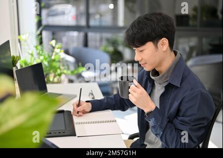 Attraktiver und kluger junger asiatischer Büroangestellter, der sich auf seine Aufgabe konzentriert, eine Liste auf seinem Notizblock erstellt, während er morgens Kaffee an seinem Schreibtisch trinkt. SI Stockfoto
