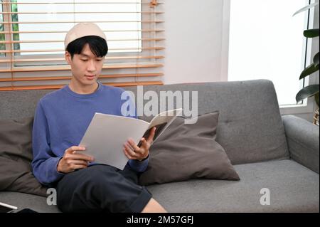 Glücklicher und entspannter junger asiatischer Mann in legerer Kleidung liest Zeitschrift, während er auf dem Sofa in seinem Wohnzimmer chillt. Stockfoto
