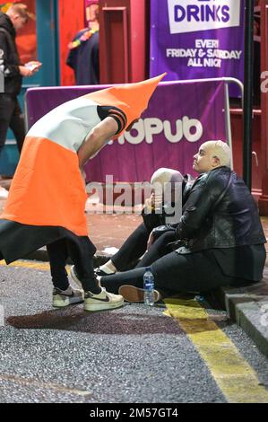 King Street, Wigan, 27. 2022. Dezember. - Reveller stolperten nach Hause und ruhten sich gegen Gebäude und auf Gehwegen aus, nach einem schönen Abend am Boxing Day, der alljährlichen Fancy Dress Night in Wigan. Ein paar Verkehrskegel stellten sicher, dass es einer Frau gut ging, als sie auf einem Bordstein saß. Ein Mann in einem Frauenkleid, Freddie Mercury-Stil, wurde angeschrien und von einer anderen Frau außerhalb von Popworld weggestoßen. Einige nahmen sich ein Essen mit, um die Nacht davor zu genießen, als sie auf Taxis warteten. Stockfoto