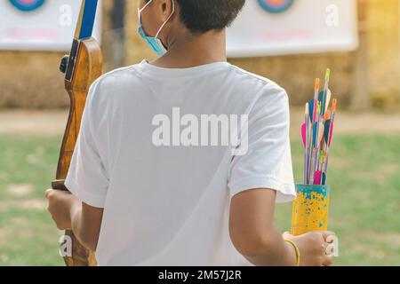 Rückansicht der asiatischen Jungen tragen Gesichtsmaske zielt Bogenschießen Bogen und Pfeil zu bunten Ziel im Schießstand während des Trainings und des Wettbewerbs. Übung und Co Stockfoto