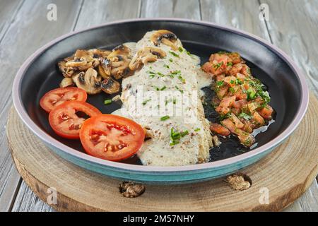 Wolfsbarsch mit Senf, Pilzen und Tomaten auf schwarzem Teller Stockfoto