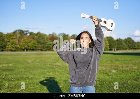 Fröhliche junge Frau, die mit ihrem Musikinstrument tanzt. Mädchen hebt ihre Ukulele hoch und posiert im Park auf grünem Feld Stockfoto