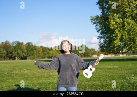 Fröhliche junge Frau, die mit ihrem Musikinstrument tanzt. Mädchen hebt ihre Ukulele hoch und posiert im Park auf grünem Feld Stockfoto