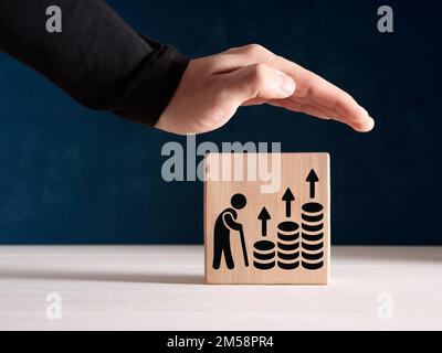 Pensionsrente, Deckungsvorsorge und Unterstützung für Ruheständler. Männliche Hand steht protektiv über dem Holzwürfel mit dem Rentner und Ascen Stockfoto