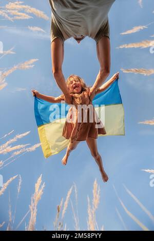 Ein Mann wirft ein kleines Mädchen auf, das die blaue gelbe Flagge der Ukraine in Händen hält, vor dem Hintergrund des klaren Himmels. Das lang erwartete Treffen von Vater und Tochter Stockfoto