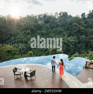 Luftaufnahme eines jungen Mannes und einer jungen Frau, die Händchen halten, während sie am Pool stehen. Ein Paar, das sich in der Nähe des Ferienresorts am Pool verliebt hat. Stockfoto