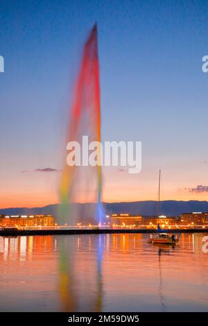 Der beleuchtete Jet d'Eau in der Abenddämmerung ist das Wahrzeichen im Genfer Seebecken, mit Segelbooten im Vordergrund und der nächtlich beleuchteten Stockfoto