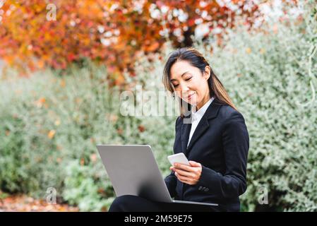 Selbstbewusste Geschäftsfrau, die mit Laptop und Smartphone arbeitet, erfolgreiche asiatische leitende Geschäftsfrau auf einer Pause im Park des Stadtteils Downton, T. Stockfoto