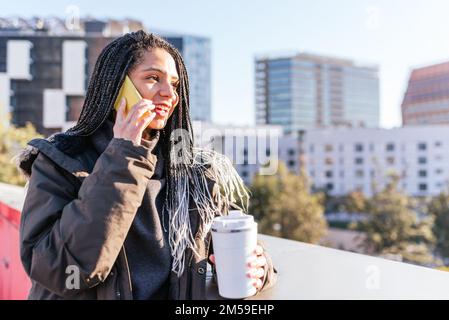 Befriedigt eine hispanische Frau mit Dreadlocks, die telefoniert, während sie in der Nähe der Grenze steht, mit einer Tasse heißem Kaffee aus der weißen Thermoskanne in der Stadt Stockfoto