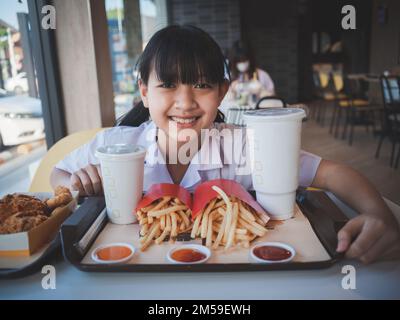 Ein süßes asiatisches Mädchen, das ein Fast Food-Set mit Pommes, Getränk und gebratenem Hähnchen in einem Café isst. Fastfood und gesundes Essen für Kinder Konzept Stockfoto