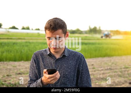 Unschärfe-Mann mit Telefon bei Sonnenuntergang. Junglandwirte, die auf einem Weizenfeld arbeiten und die Bewässerungszentrale mit Drehsprengesystem per Smartphone überprüfen und einstellen. Stockfoto