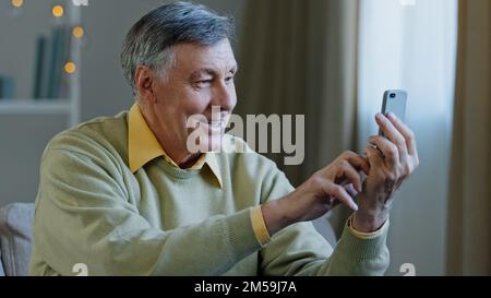 Glücklicher älterer erwachsener Mann, der ein Telefon hält, führen Sie ein Videogespräch mit einem lächelnden Großvater, der auf den Smartphone-Bildschirm schaut, kommunizieren Sie mit einem modernen Gerät Stockfoto