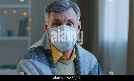 Nahaufnahme Erwachsener reifer Mann trägt medizinische Maske Blick auf Kamera krank alter Mann sitzt drinnen in einer Decke Porträt ernsthaft ältere Menschen pensioniert Pandemie Stockfoto